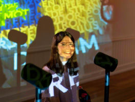 A girl sitting with a standing microphone in front of her and brightly colored letters and words projected on her face