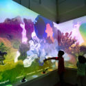 Two children in the Word Worlds gallery. They are backlit, and the wall behind them is a vibrant swirl of colors with descriptive words like "tempestuous," "crepuscular," and "magical" at the bottom.