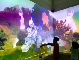 Two children in the Word Worlds gallery. They are backlit, and the wall behind them is a vibrant swirl of colors with descriptive words like "tempestuous," "crepuscular," and "magical" at the bottom.