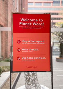 A sign with COVID-19 safety guidelines for visitors to Planet Word. Text reads: Welcome to Planet Word! Your journey with words starts with these words right here— please follow our COVID-19 safety guidelines: Stay 6 feet apart. (What has 6 feet and three eyes? Social Distancing.) Wear a mask. (Everyone loves a mystery.) Use hand sanitizer. (Lady Macbeth would approve.) Thank You!