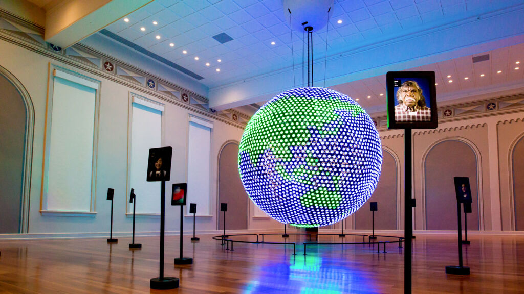 A large blue and green LED globe in Planet Word's Spoken World gallery showing the Asian continent and Oceania. Standing tablets are placed around the room.